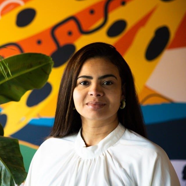 Luana, founder of Baioca, smiling in front of a colourful, tropically painted wall with large green plants.