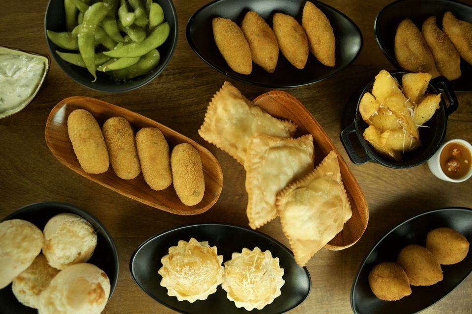 Many bowls of petiscos seen from above: coxinhas, pastéis, pão de queijo, edamame and tartlets.